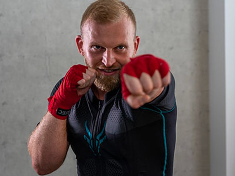 Christoph Rüsseler beneficia dell'allenamento EMS con Antelope durante la kickboxing. Esegue un movimento di pugno e guarda intensamente la telecamera. Indossa bende rosse da boxe sulle mani. 